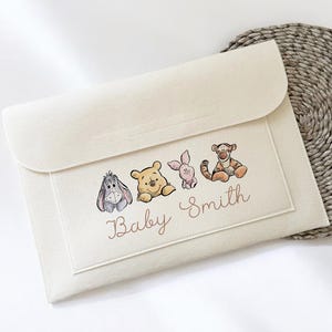May include: A cream-coloured felt baby clutch bag with a flap closure. The front displays illustrations of Winnie the Pooh characters: Eeyore, Winnie the Pooh, Piglet, and Tigger. The text "Baby Smith" is written below the characters.