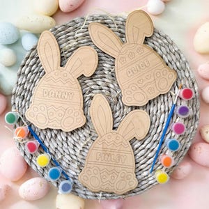 May include: Three unfinished wooden bunny-shaped Easter egg ornaments with the names Alice, Danny, and Finley. The ornaments are on a woven basket surface with paint pots and brushes.
