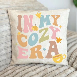 May include: A beige throw pillow with the text "In My Cozy Era" in colourful, retro-style lettering. The pillow features a small pumpkin, a cup of coffee, and autumn leaves.