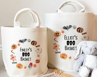 Può includere: Due cesti in tela color bianco sporco con manici, ciascuno caratterizzato da una grafica circolare di icone di Halloween tra cui zucche, un pipistrello, un fantasma e un omino di pan di zenzero. Il cesto a sinistra riporta Finley's BOO BASKET.