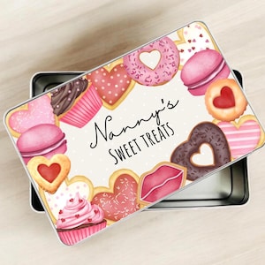 May include: A rectangular metal tin with a lid decorated with illustrations of pink and brown sweets, such as heart-shaped biscuits, cupcakes, and macarons. The lid has the text "Nanny's SWEET TREATS" in cursive.