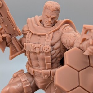 May include: A brown action figure of a muscular character in a combat pose, holding a large gun and a shield. The figure has detailed armor, pouches, and a determined facial expression. The figure is approximately 6 inches tall.