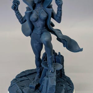 Peut inclure: Figurine bleue d'un personnage féminin tenant deux pistolets, vêtue d'un manteau et de bottes. La figurine est posée sur une base rocheuse avec une bordure circulaire blanche. Le personnage a des oreilles pointues et une expression déterminée.