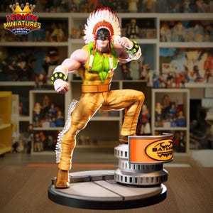 May include: A detailed figurine of a character in a dynamic pose, wearing a headdress, mask, and yellow and green outfit. The figure stands on a platform with the Batman logo. The base is black and silver.