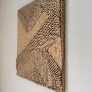 May include: A square, textured wall art piece with a geometric design. The artwork is constructed from natural-colored rope, creating a layered effect with varying shades of tan and brown. The piece is mounted on a wooden frame.