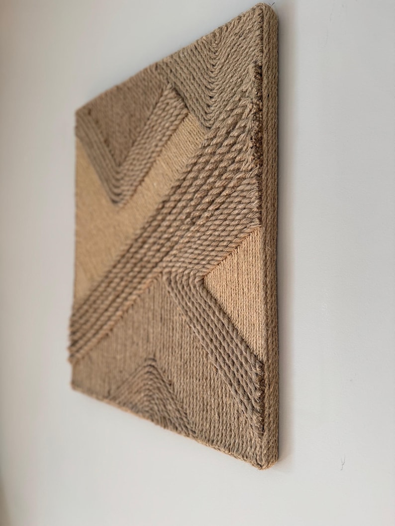 May include: A square, textured wall art piece with a geometric design. The artwork is constructed from natural-colored rope, creating a layered effect with varying shades of tan and brown. The piece is mounted on a wooden frame.