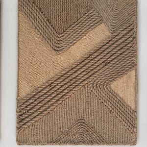 May include: A rectangular wall art piece with a geometric design made from natural rope. The design features diagonal and angular lines in varying shades of brown and tan, creating a textured, three-dimensional effect.