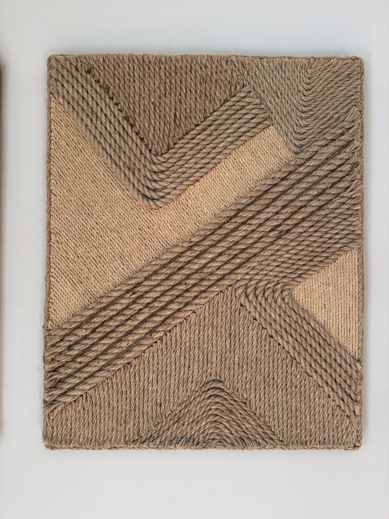 May include: A rectangular wall art piece with a geometric design made from natural rope. The design features diagonal and angular lines in varying shades of brown and tan, creating a textured, three-dimensional effect.