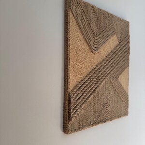 May include: A square, textured wall art piece with a geometric design. The artwork features a natural, beige color palette with rope detailing. The design includes diagonal and zig-zag patterns, creating a modern, tactile aesthetic.