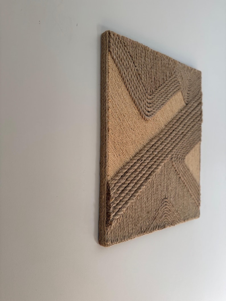 May include: A square, textured wall art piece with a geometric design. The artwork features a natural, beige color palette with rope detailing. The design includes diagonal and zig-zag patterns, creating a modern, tactile aesthetic.