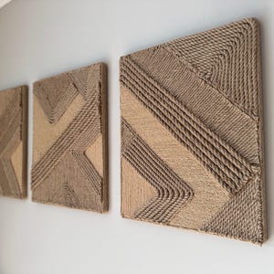 May include: Three rectangular wall art pieces with geometric designs made from natural-colored rope. The artwork is mounted on a light gray wall, showcasing a modern, textured aesthetic. The rope is tightly woven, creating a tactile, dimensional effect.