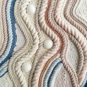Op de afbeelding: Getextureerde wandophanging met een geweven ontwerp. Het stuk heeft een palet van zachte kleuren, waaronder crème, lichtblauw en perzik, met drie witte bolvormige accenten. Het ontwerp bevat touwachtige texturen en gebogen lijnen.