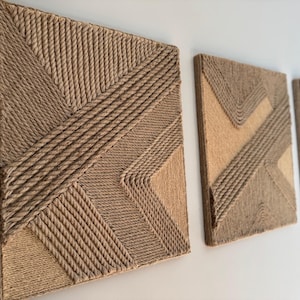 May include: Three rectangular wall art pieces with geometric designs made from woven natural fiber rope. The artwork features a mix of diagonal and straight lines in shades of tan and brown, creating a textured, modern aesthetic.
