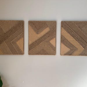 May include: Three rectangular wall art pieces with geometric designs. The artwork features a combination of light and dark brown rope, creating textured patterns. The pieces are mounted on a white wall, adding a natural, neutral aesthetic.