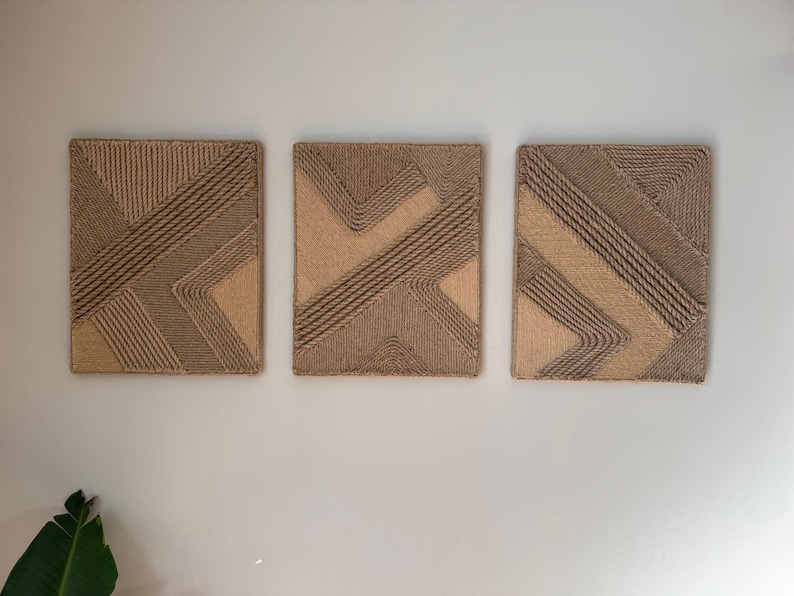 May include: Three rectangular wall art pieces with geometric designs. The artwork features a combination of light and dark brown rope, creating textured patterns. The pieces are mounted on a white wall, adding a natural, neutral aesthetic.