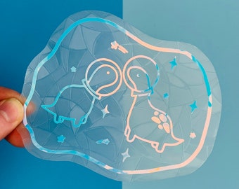 May include: A clear sticker with a holographic finish featuring two dinosaurs wearing astronaut helmets. The dinosaurs are facing each other and are surrounded by stars.