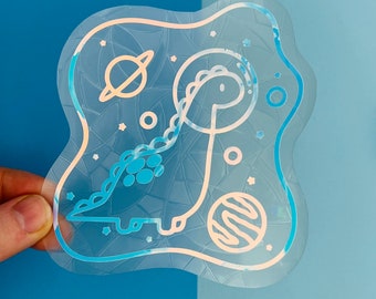 May include: A holographic sticker featuring a cartoon dinosaur in a spacesuit with planets and stars in the background.