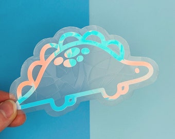 Könnte beinhalten: Ein transparenter, schimmernder Aufkleber mit einem Cartoon-Dinosaurier mit einem rosa-blauen Wirbelmuster. Der Dinosaurier ist blau umrandet und hat einen rosa Bauch.