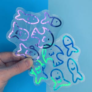 May include: Two clear, iridescent stickers featuring fish designs. One sticker has purple outlines, the other has blue and green outlines. The fish have simple, cartoonish features. The stickers are held against a blue background.