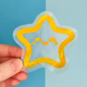 May include: A clear sticker with a yellow star shape. The star has a happy face with a small smile. The sticker is on a blue background.