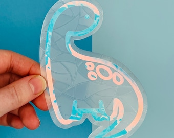 May include: A clear iridescent sticker of a cartoon dinosaur with a pink outline. The sticker has a blue and white marbled pattern.