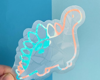 May include: A clear iridescent sticker of a cartoon dinosaur with a teal and orange outline. The dinosaur has a spiky back and a long neck.