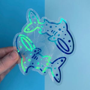 May include: A clear holographic sticker with two shark designs. The sharks are outlined in blue and green, with dotted patterns. The sticker is held against a backdrop of light and dark blue.