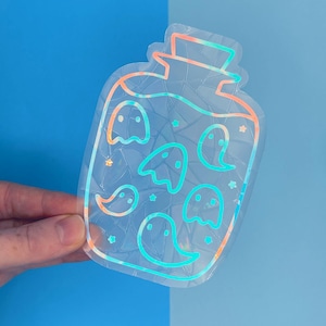 May include: A clear sticker with a jar design containing cartoon ghosts. The sticker has a holographic finish, with the jar and ghost outlines in orange, blue, and green. The background is a gradient of blue.