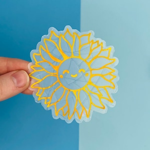May include: A clear sticker with a gold outline of a smiling sunflower. The sunflower has a simple face with two eyes and a smile.
