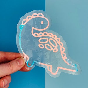 May include: A clear, iridescent dinosaur sticker with a pink and blue outline. The sticker has a geometric pattern inside the dinosaur shape and is held against a blue background. The dinosaur has a friendly, cartoonish design.