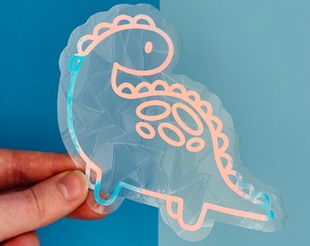 Pode incluir: Um autocolante de dinossauro transparente e iridescente com um contorno rosa e azul. O autocolante apresenta um padrão geométrico dentro da forma do dinossauro e é mantido contra um fundo azul. O dinossauro tem um design amigável e de desenho animado.