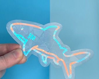 May include: A holographic sticker of a shark with a blue and orange iridescent finish. The sticker is outlined in white.