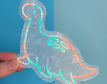 Könnte beinhalten: Ein transparenter, schimmernder Aufkleber mit einem Cartoon-Dinosaurier mit türkisfarbenem Umriss. Der Aufkleber hat eine holografische Oberfläche.