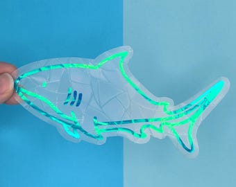 May include: A clear iridescent sticker of a shark with a teal outline. The sticker is on a blue background.