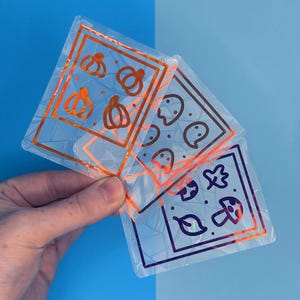 May include: Transparent, square stickers with orange and purple borders. The stickers display autumn-themed illustrations of pumpkins, ghosts, leaves, and mushrooms. The stickers are held against a blue backdrop.