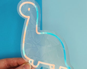 Pode incluir: Um adesivo transparente iridescente de um dinossauro de desenho animado com contorno branco. O adesivo tem um padrão texturizado e espiral.