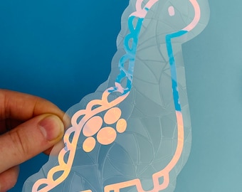 Könnte beinhalten: Ein holografischer Aufkleber mit einem hellblauen Dinosaurier mit rosa Umrandung. Der Dinosaurier hat ein weißes Auge und einen weißen Fleck auf dem Rücken.