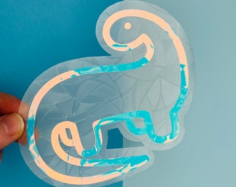 May include: A holographic sticker of a blue dinosaur with a white outline. The sticker has a textured, iridescent finish.