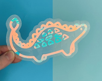 Pode incluir: Um adesivo holográfico de um dinossauro turquesa e laranja com uma espinha dorsal espinhosa.