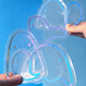 May include: Clear, iridescent stickers with a whimsical design. The stickers have a layered effect with blue, purple, and pink outlines. The stickers are held against a blue background. The design appears to be a stylized cloud or abstract shape.
