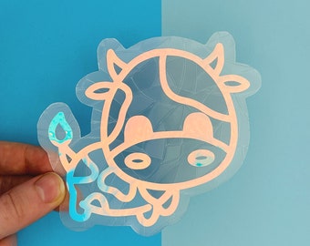 May include: A holographic sticker of a cartoon cow with a pink outline. The cow is facing the right side of the image and has a single udder.