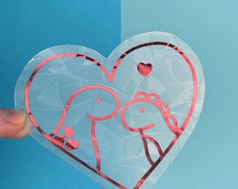 May include: A clear heart-shaped sticker with a pink outline featuring two cartoon dinosaurs with a small pink heart above them.