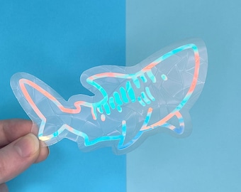 May include: A clear iridescent sticker of a shark outline. The sticker has a holographic finish that shifts colors from blue to pink.
