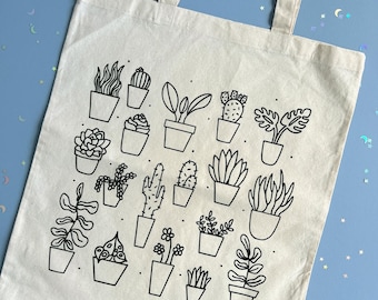 May include: A white canvas tote bag with a black line drawing of potted plants. The plants include succulents, cacti, and other leafy plants.