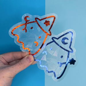 May include: Two clear stickers with a cracked ice texture. One sticker has orange outlines, the other has blue outlines. Both have cartoon-like faces and are held against a blue background.