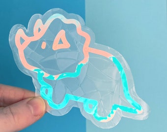 May include: A clear iridescent sticker of a cartoon triceratops with a pink outline. The sticker has a textured, cracked glass effect.