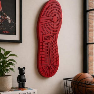 Peut inclure: Décoration murale en forme de semelle de chaussure de sport rouge avec le texte "DRE'S". L'art mural est monté sur un mur de couleur claire. Une photo encadrée d'un joueur de basket-ball et un ballon de basket sont également visibles.
