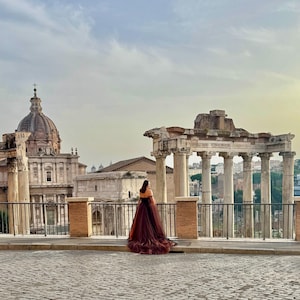 May include: A person in a long, flowing burgundy dress stands on a cobblestone street, facing away from the camera. Ancient Roman ruins and a domed building are in the background under a cloudy sky.