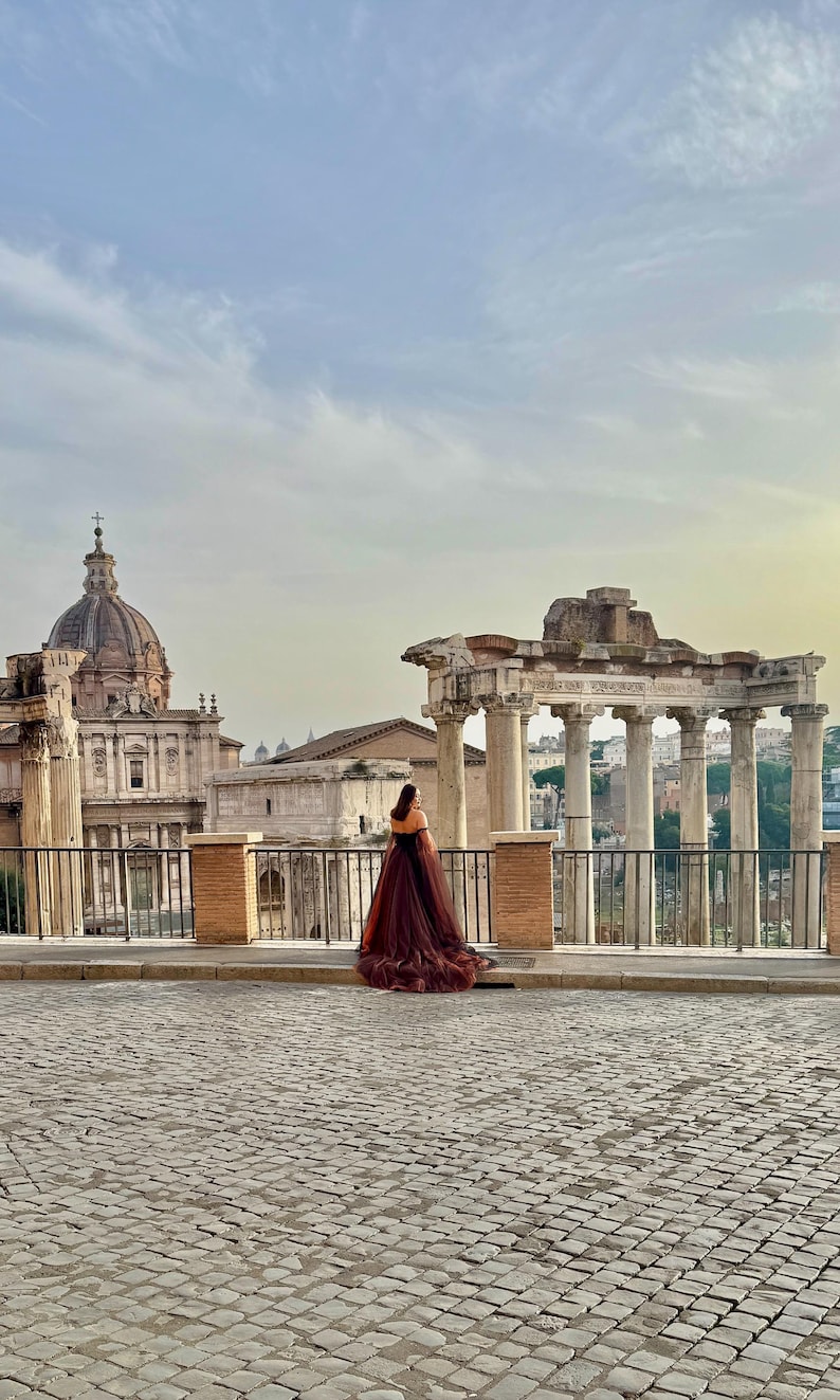 May include: A person in a long, flowing burgundy dress stands on a cobblestone street, facing away from the camera. Ancient Roman ruins and a domed building are in the background under a cloudy sky.