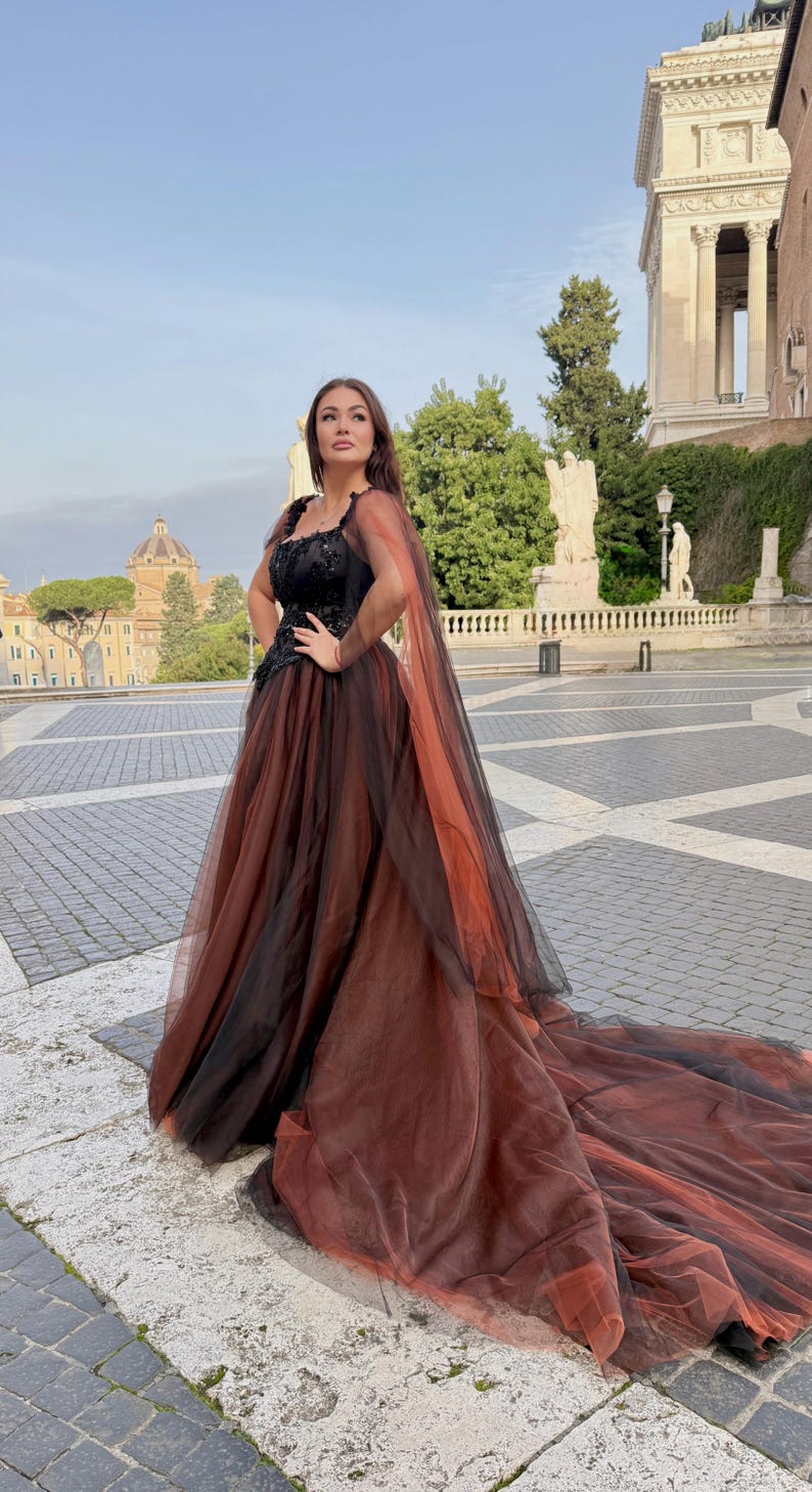 May include: A formal gown with a black bodice and a flowing skirt that transitions from brown to black. The dress features a long train and sheer cape. The woman is standing outdoors in a European city square.